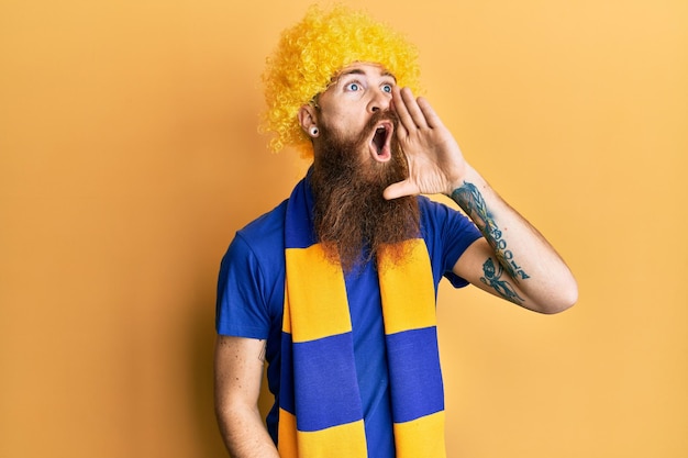 Gratis foto roodharige man met lange baard voetbal hooligan juichen spel dragen grappige pruik schreeuwen en schreeuwen luid naar de andere kant met de hand op de mond communicatieconcept