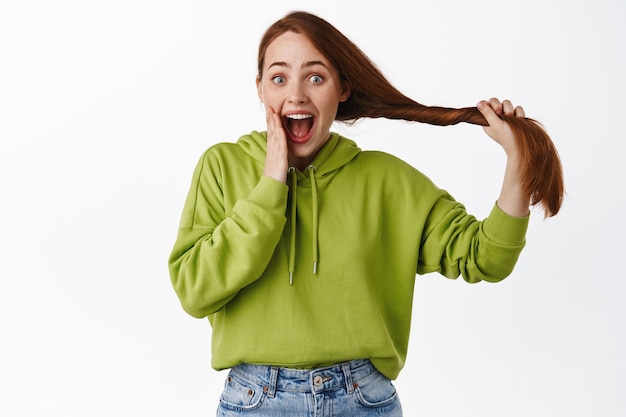 Roodharige meid ziet er opgewonden uit met haarverzorgingsbehandeling, houdt sterk natuurlijk rood haar vast en kijkt verrast en verbaasd op wit