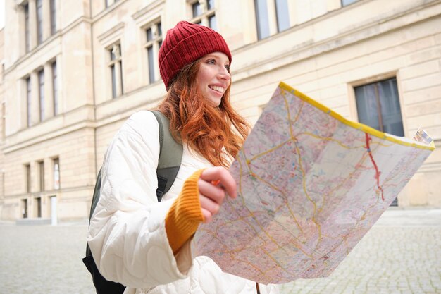 Gratis foto roodharige meisjestoerist verkent de stad, kijkt naar de papieren kaart om een weg te vinden voor historische bezienswaardigheid