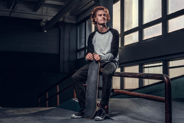 Roodharige skateboarder die zijn bord vasthoudt terwijl hij op een grindrail in het skatepark binnenshuis zit.