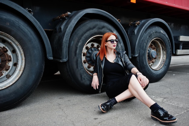 Roodharige stijlvolle meisjeskleding in het zwart zittend tegen grote vrachtwagenwielen
