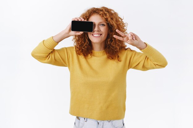 roodharige vrouw met krullend haar in gele trui, smartphone horizontaal houdend en haar ogen bedekkend met mobiele telefoon