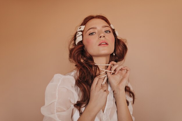 Roodharige vrouw met roze wangen speelt met haar parelketting. Dame in klassieke witte blouse die neer camera bekijkt.