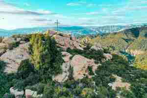 Gratis foto rotsachtige klif bedekt met groen met een kruis op de top en prachtige bergen