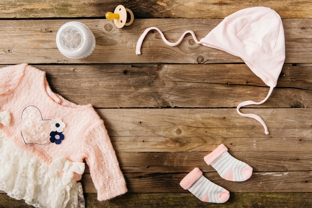 Roze babyjurk met hoofddeksels; paar sokken; melkfles en fopspeen op houten tafel