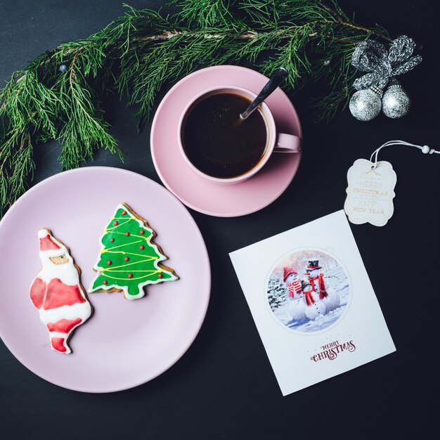 Roze kop koffie, bord met kerstmisbroodjes en briefkaart liggen op zwarte tafel