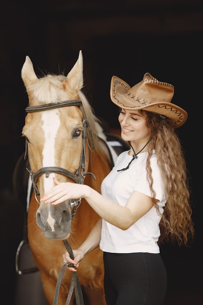 Ruitervrouw die aan haar paard in een stal spreekt. Vrouw heeft lang haar en wit t-shirt. Achtergrond is donker en zwart.