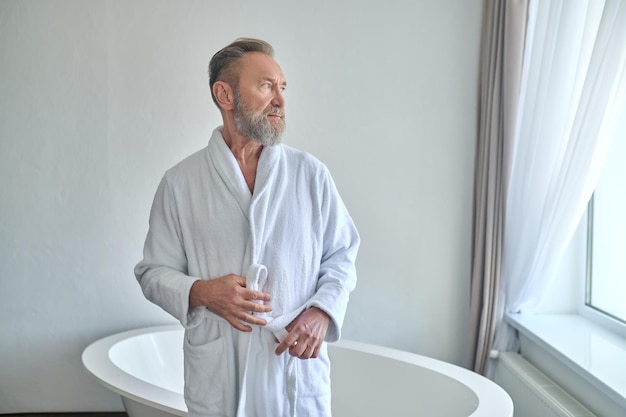 Rustige nadenkende grijsharige bebaarde volwassen man met snor die de riem van zijn witte badstof badjas vastmaakt