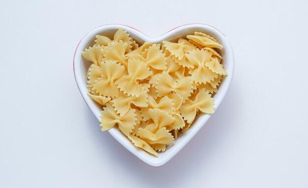 Ruwe farfalle pasta in een hartvormige kom op witte muur, bovenaanzicht.