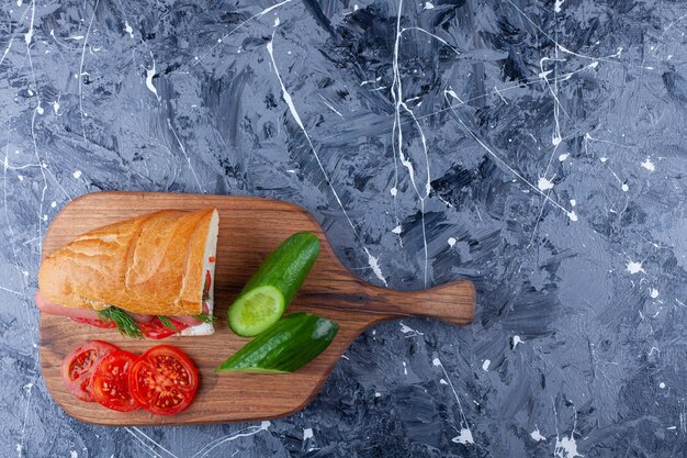 Sandwich, gesneden komkommer en tomaten op snijplank op blauw.