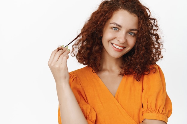 Sassy en mooie roodharige vrouw met krullend kort haar, rolkrul op vinger, giechelen en koket glimlachen, flirten, glimlachen verleidelijk, staande op wit