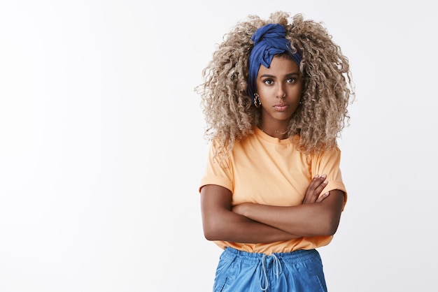 Sassy en vastberaden knap stijlvol afro-amerikaans blond meisje afro kapsel in trendy hoofdband oranje tshirt kruis armen borst kijken camera serieus en cool staande witte achtergrond