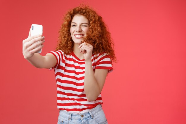 Sassy knappe stijlvolle charismatische roodharige vrouwelijke krullend kapsel knipogen brutale uitdrukking maken flirterige kinky gezichten houden smartphone nemen selfie opnemen videoboodschap spelen grappige gezichtsfilters.