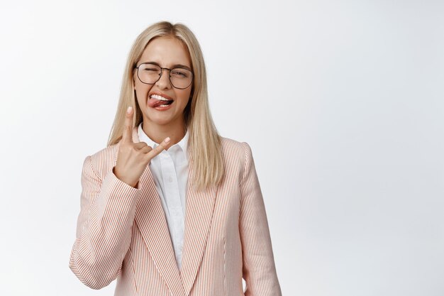 Sassy zakenvrouw die tong en rock heavy metal gebaar toont met plezier op het werk in pak tegen een witte achtergrond