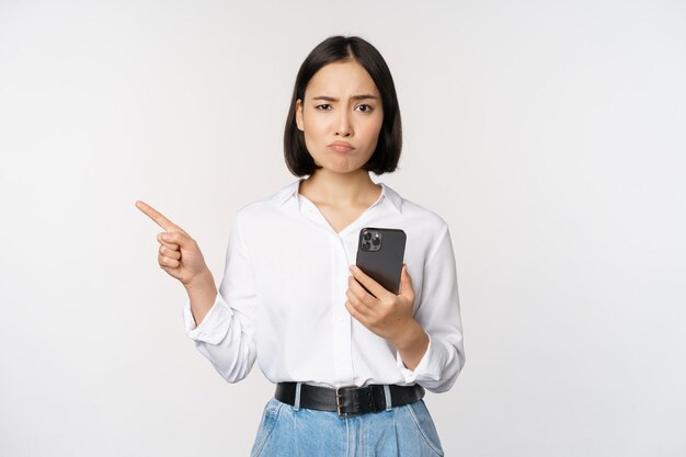 Sceptisch Aziatisch modern meisje met smartphone wijzende vinger links naar lege ruimte en fronsend aarzelend twijfelt over witte achtergrond