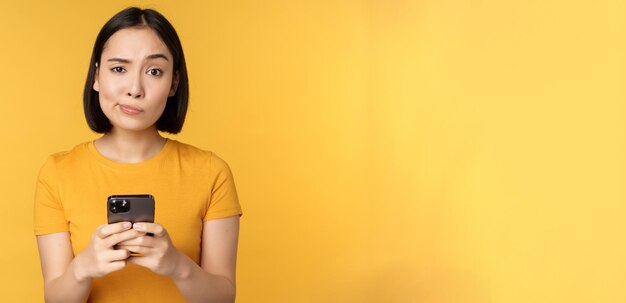 Sceptische aziatische vrouw die een smartphone vasthoudt en met twijfel naar de camera kijkt die over een gele achtergrond staat