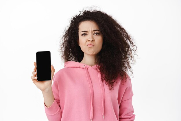 Sceptische brunette vrouw die klaagt over een leeg mobiel scherm en grimassen, teleurgesteld door de smartphone-app die in een roze hoodie tegen een witte achtergrond staat