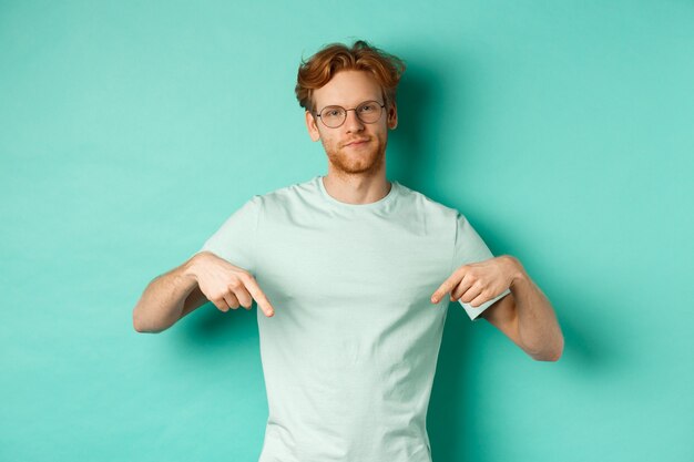 Sceptische en lastige man met rood haar en baard, bril en t-shirt, grijns en wijs met de vingers naar beneden, met promo met oordeelkundig gezicht, turkooizen achtergrond.