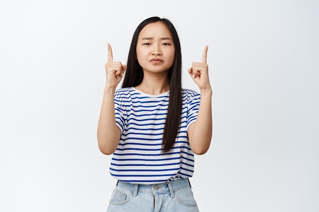 Sceptische jonge aziatische vrouw heeft twijfels, fronst en kijkt terughoudend, wijst vingers omhoog op wit.