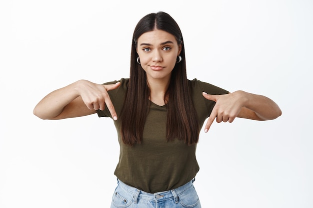 Sceptische jonge brunette vrouw die twijfels heeft, vingers naar beneden wijst naar iets slechts of onaangenaams, staande tegen een witte muur