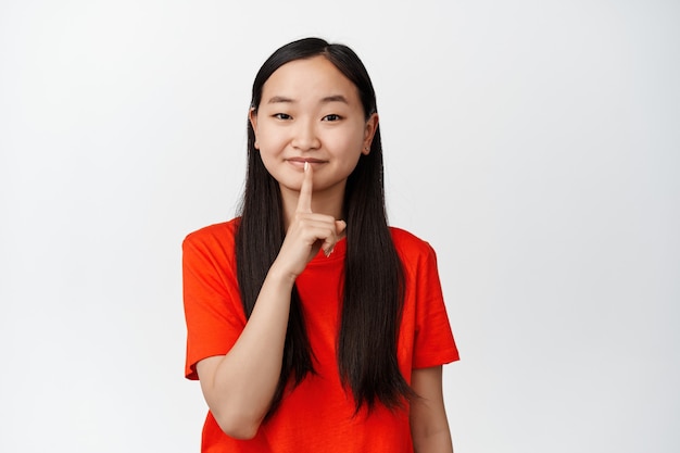 Schattig aziatisch meisje lacht en zwijgt, toont een shhh hush-teken, zegt stil te zijn, staat in een rood t-shirt op wit