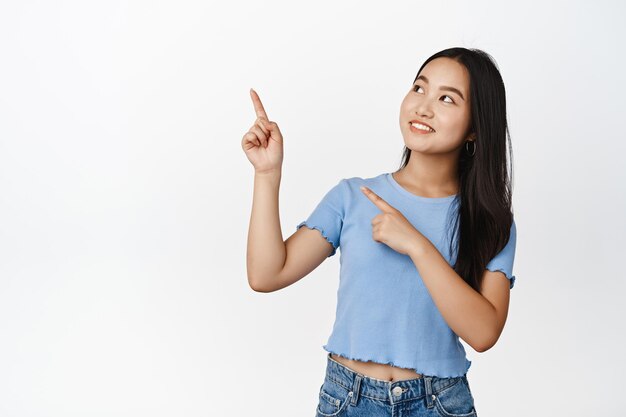 Schattig Aziatisch meisje wijzend en kijkend naar de linkerbovenhoek met een geïntrigeerde glimlach die tekst leest op een lege ruimte witte achtergrond