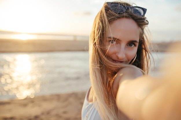 Schattig gelooid meisje met steil haar selfie maken op natuur achtergrond Knappe vrouw in zonnebril nemen foto van zichzelf terwijl staande aan de zeekust vroeg in de avond