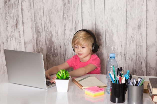 schattig kind jongen in roze t-shirt zwarte koptelefoon met behulp van grijze laptop op tafel samen met groene plant fles pennen op de grijze muur