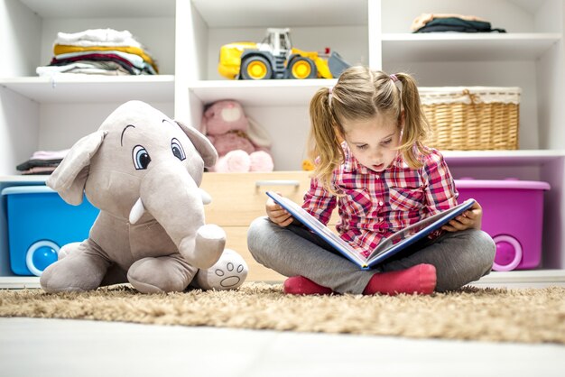 Schattig klein meisje dat op het tapijt zit en een boek leest voor haar knuffelolifant