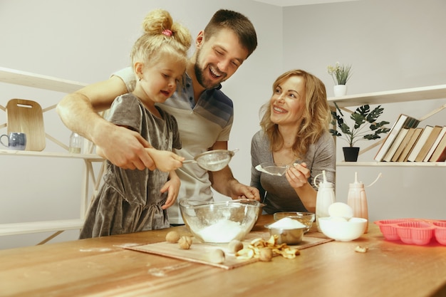 Schattig klein meisje en haar mooie ouders bereiden het deeg voor de taart in de keuken thuis.