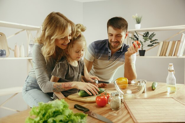 Schattig klein meisje en haar mooie ouders zijn groenten snijden en glimlachen tijdens het maken van salade in de keuken thuis. Familie levensstijl concept
