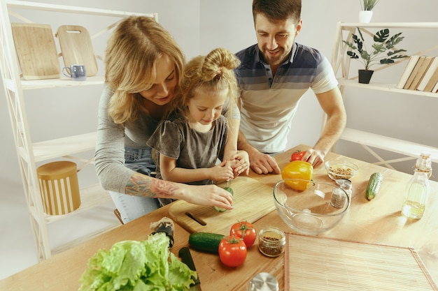 Schattig klein meisje en haar mooie ouders zijn groenten snijden en glimlachen tijdens het maken van salade in de keuken thuis. Familie levensstijl concept