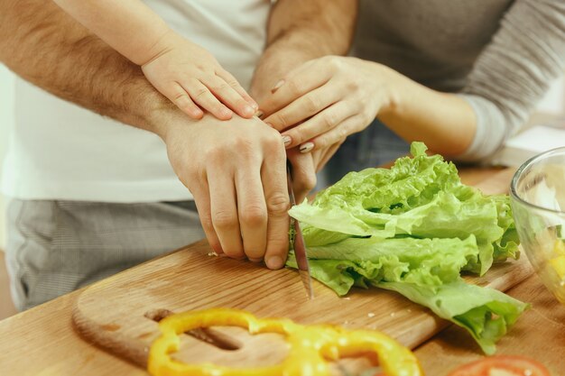 Schattig klein meisje en haar mooie ouders zijn groenten snijden en glimlachen tijdens het maken van salade in de keuken thuis