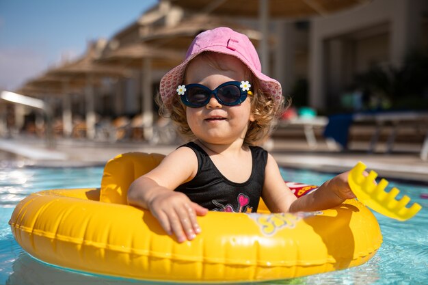 Schattig klein meisje in een hoed en zonnebril speelt in het zwembad zittend in een cirkel zwemmen