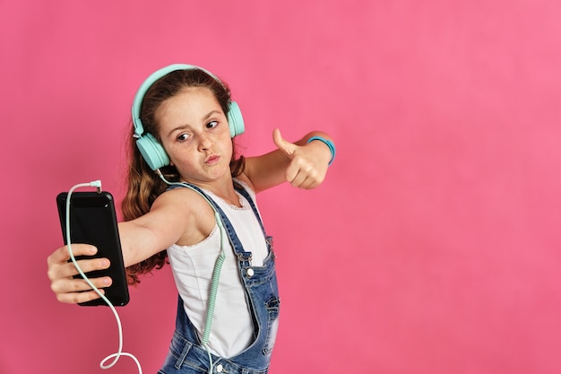 Schattig klein meisje, luisteren naar muziek met een telefoon en koptelefoon op een roze muur