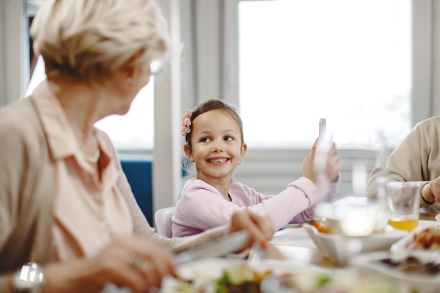 Gratis foto schattig klein meisje met behulp van slimme telefoon en communiceren met haar grootmoeder tijdens een maaltijd aan de eettafel