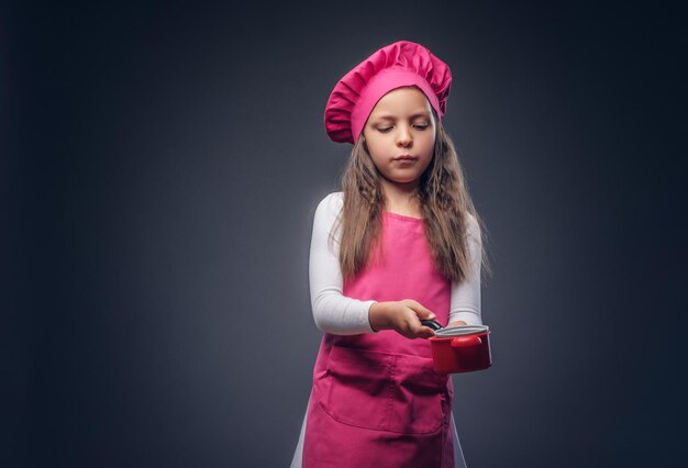 Schattig mooi schoolmeisje gekleed in een roze kok uniform houdt kookgerei in een studio. Geïsoleerd op een donkere achtergrond.