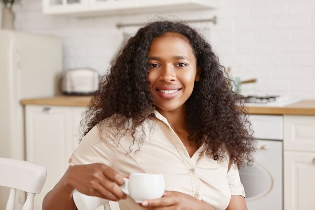Schattig positief twintig-jarige mulat meisje met volumineuze Afro kapsel met vrolijke gelukkige gezichtsuitdrukking, genieten van mooie ochtend thuis, zittend in de keuken, breed glimlachend