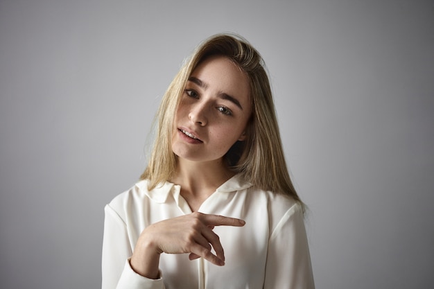 Schattig schattig blond meisje in witte blouse poseren geïsoleerd in studio, glimlachend in de camera. Mooie stijlvolle jonge vrouw, gekleed in zijdeachtig elegant shirt, kantelend hoofd, wijsvinger zijwaarts wijzend