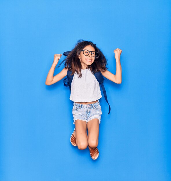 Schattig Spaans studentkindmeisje met bril en rugzak die gelukkig glimlacht Springend met een glimlach op het gezicht over een geïsoleerde blauwe achtergrond