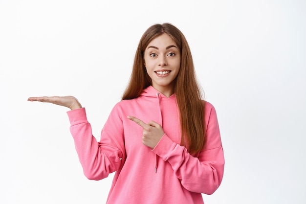 Schattig tienermeisje wijst met de vinger naar de lege hand, toont het copyspace-logo op haar handpalm, glimlacht naar voren en staat over een witte muur