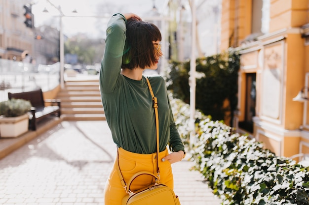 Schattig vrouwelijk model draagt trendy handtas ontspannen tijdens wandeling. Buiten schot van betoverende kortharige brunette vrouw rondkijken op straat.