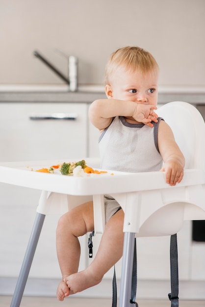Schattige babyjongen in kinderstoel groenten eten in de keuken