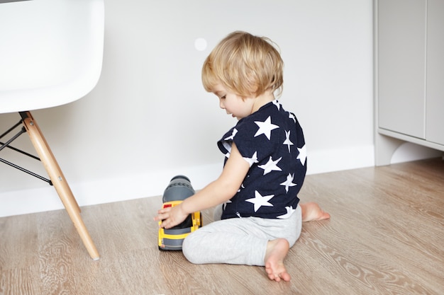 Schattige babyjongen met blond haar zittend op een houten vloer in zijn slaapkamer, zijn favoriete speeltje te houden en glimlachen. Peuter plezier, spelen met gele plastic vrachtwagen. Vroeg leren.