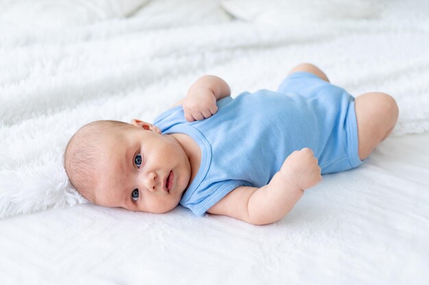 Schattige babyjongen van drie maanden oud in een blauwe bodysuit op een wit bed thuis