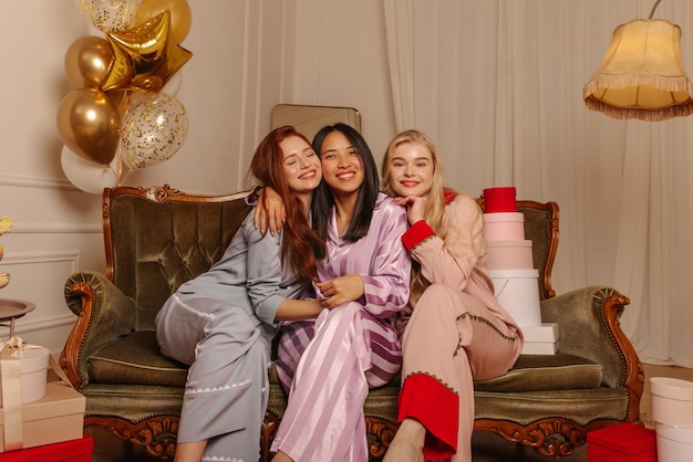Schattige blanke jonge meisjes in pyjama's kijken naar de camera, zitten knuffelend op de bank en vieren vrijgezellenfeest Rusttijdconcept