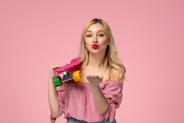 Schattige blonde meid met rode lippenstift in roze blouse die kus met skateboard verzendt