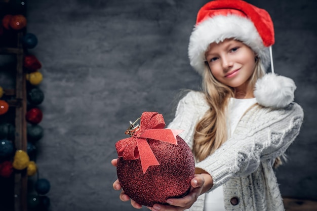 Schattige blonde meisje in de hoed van de Kerstman houdt rode kerstbal.