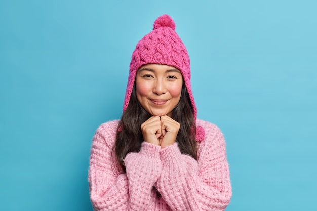 Schattige brunette Aziatisch meisje houdt handen onder kin kijkt aangenaam aan voorzijde draagt winter hoed en trui klaar voor wandeling tijdens koude dag geïsoleerd over blauwe muur