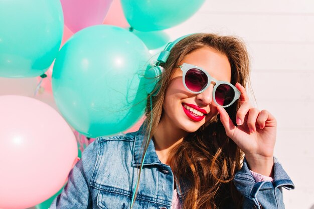 Schattige brunette jonge vrouw kijkt door een stijlvolle zonnebril en poseert met een glimlach naast kleurrijke ballonnen. Close-up portret van blij feestvarken dat muziek luistert in turquoise koptelefoon